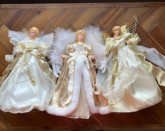 May include: Three decorative angel figurines with blonde hair and white and gold gowns. Each angel has feathered wings and is posed on a wooden surface. The angels are a festive holiday decoration.