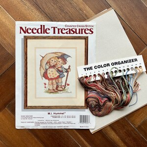 May include: A counted cross stitch kit featuring a vintage illustration of two children under an umbrella. The kit includes a color organizer with floss, Aida cloth, and instructions. The kit is titled "Sunny Weather" and is by M.I. Hummel.