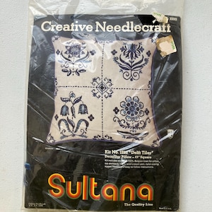 May include: A sealed package for a Creative Needlecraft kit. The kit features a pillow design with Delft tiles, a 13-inch square pattern with blue floral motifs on a cream background. The package includes the Sultana brand logo.