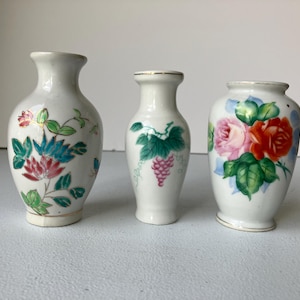May include: Three small, white ceramic vases with hand-painted floral designs. The vases feature pink, red, and blue flowers, green leaves, and a bunch of grapes. Each vase has a unique shape and design.