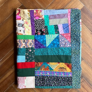May include: A handmade patchwork quilt with a variety of colorful fabric squares. The quilt features a mix of floral, geometric, and solid patterns in shades of green, blue, purple, red, and yellow. The quilt is rectangular and appears to be folded.