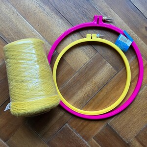 May include: A bundle of yellow yarn wrapped in clear plastic, next to two embroidery hoops. One hoop is bright pink, the other is yellow. The hoops are different sizes, and the background is a wooden floor.