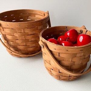 Op de afbeelding: Twee bruine gevlochten manden met houten handvatten. De kleinere mand is gevuld met rode appels.