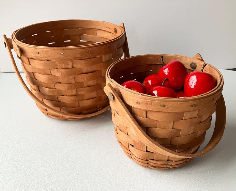 Op de afbeelding: Twee bruine gevlochten manden met houten handvatten. De kleinere mand is gevuld met rode appels.