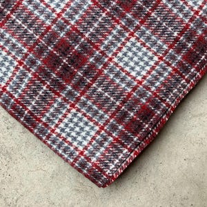 May include: Close-up of a folded fabric with a classic plaid pattern. The fabric features a combination of red, white, and gray hues, creating a visually appealing design. The texture of the fabric appears soft and possibly woolen.