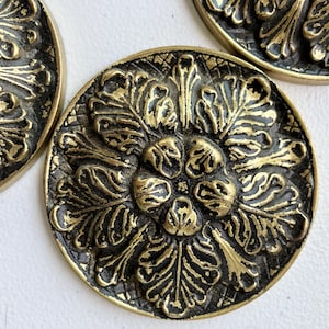 May include: Close-up of several antique brass buttons with an ornate floral design. The buttons are round with a detailed pattern of leaves and a central flower. The buttons have a dark, aged finish, suggesting vintage or retro style.