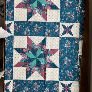 May include: A teal blue fabric with a patchwork design featuring a repeating pattern of pink, green, and white stars. The fabric has a white background with small white dots.