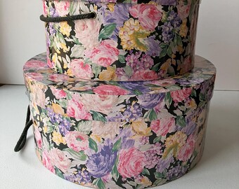 May include: Two round hat boxes with floral patterns. The top box has a black rope handle. Both boxes have a black background with pink, purple, and yellow flowers.