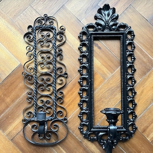 May include: Two black metal candle holders with ornate designs. The holder on the left has a more intricate design with swirling details. The holder on the right has a more rectangular frame with a decorative top.