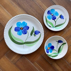 May include: A set of three white ceramic plates with a hand-painted floral design. Each plate features a blue flower with green leaves and stems. The plates vary in size, with the largest measuring approximately 10 inches in diameter.