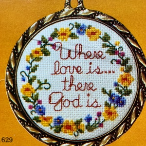 May include: A round, white cross-stitch embroidery with a gold frame. The embroidery features a wreath of yellow and blue flowers with green leaves and the text "Where love is there God is".