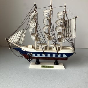 May include: A miniature wooden sailboat model with white sails and a blue and white hull. The model has black rigging and is mounted on a white base with the word "BELEM" printed on it. The ship has an anchor.