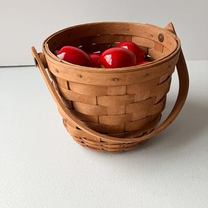 Op de afbeelding: Een kleine, houten mand met een handvat, gevuld met rode appels. De mand is gemaakt van geweven hout en heeft een natuurlijke afwerking.