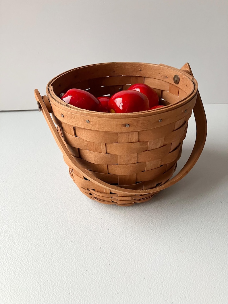 Op de afbeelding: Een kleine, houten mand met een handvat, gevuld met rode appels. De mand is gemaakt van geweven hout en heeft een natuurlijke afwerking.