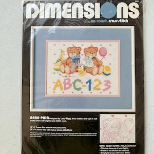 May include: A Dimensions no count cross stitch kit featuring a framed cross-stitch design of two teddy bears, ABC-123, and toys. The kit includes a 12"x9" frame size without mat (30x23cm) and a 16"x13" frame size with mat (40x33cm).