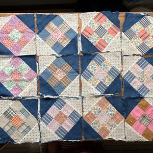 May include: A collection of twelve patchwork quilt squares. Each square features a central diamond pattern in various colors like pink, blue, and orange, surrounded by a dark blue border. The squares are arranged on a wooden surface.