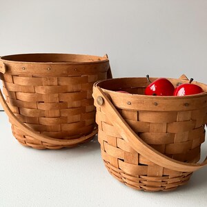 Op de afbeelding: Twee bruine gevlochten manden met handvatten. De kleinere mand bevat drie rode appels.