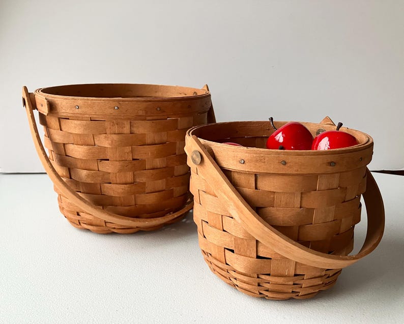 Op de afbeelding: Twee bruine gevlochten manden met handvatten. De kleinere mand bevat drie rode appels.