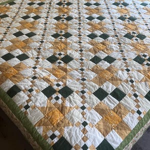 May include: A patchwork quilt featuring a repeating geometric pattern of white, mustard yellow, and dark green fabric squares. The design incorporates star-shaped motifs and a thin green border, with visible stitching throughout the textile surface.