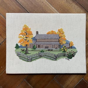 May include: Cross-stitch artwork of a cabin scene. The cabin has a gray roof and is surrounded by trees with yellow and orange leaves. A green fence is in the foreground. The artwork is on a canvas.