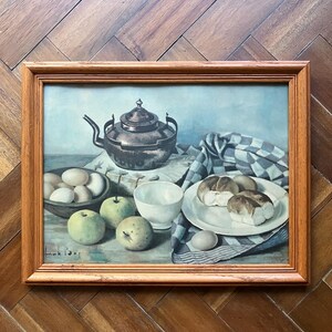 May include: A framed still life painting featuring a copper tea kettle, apples, eggs, bread rolls, and a white bowl and plate. The painting has a light blue background and a wooden frame. The painting is signed.
