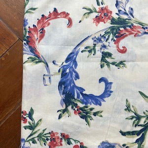 May include: Close-up of a white fabric with a repeating floral pattern. The design features red, blue, and green botanical elements. The fabric has a vintage aesthetic, with a detailed print of flowers and leaves.