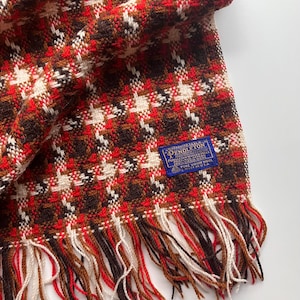 May include: A woven scarf featuring a houndstooth pattern in shades of red, brown, and cream. The scarf has a blue rectangular label with the text Pendleton, Portland, Oregon, 100% Virgin Wool, Made in U.S.A. It includes a fringed edge with red, brown, and cream threads.