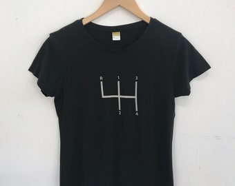 May include: Black t-shirt with a white graphic of a four-speed manual transmission gear shift pattern. The numbers 1, 2, 3, 4, and R are printed on the gear shift pattern.