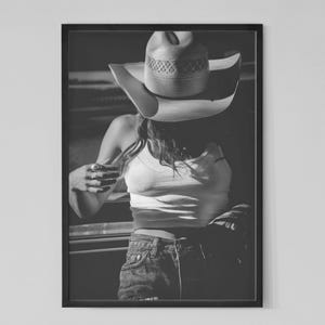 May include: Black and white framed print of a person wearing a cowboy hat, tank top, and denim shorts. The image is a portrait, with the person's face partially obscured by the hat. The overall aesthetic is vintage and evokes a sense of the American West.