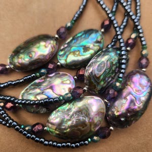 May include: A multi-strand necklace featuring iridescent abalone shell pendants. The oval-shaped pendants display a range of colors, including blues, greens, and purples. The necklace also incorporates small, dark beads and purple accent beads.