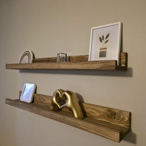 May include: Two rustic, wooden floating shelves mounted on a neutral wall. The top shelf holds a framed botanical print, a clear cube, and a ceramic rainbow. The bottom shelf displays a gold hand sculpture and a white square object.