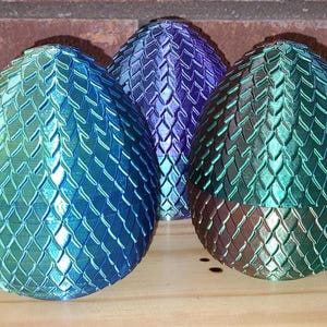 Puede incluir: Tres adornos iridiscentes en forma de huevo de dragón con textura escamosa. Los huevos son azules, morados y verdes.