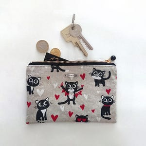 Peut inclure: Une petite pochette rectangulaire zippée avec un motif répété de chats noirs et de coeurs rouges sur un fond gris clair. La pochette a une fermeture éclair dorée et un pompon noir. Des clés et des pièces sont à côté.