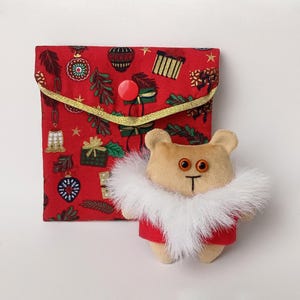 May include: A small plush brown bear wearing a red coat with a white fur collar. The bear is standing in front of a red fabric pouch with a Christmas pattern.