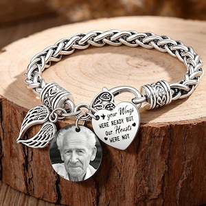 May include: A silver memorial bracelet with a braided chain. It has a round charm with a black and white photo, a heart-shaped charm with text, and a wing charm. The text on the heart charm reads: "Your Wings Were Ready But Our Hearts Were Not."
