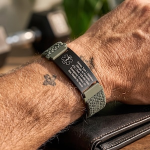 Op de afbeelding: Een olijfgroene geweven medische alarmarmband met een zwarte metalen plaat met het Star of Life-symbool en gegraveerde tekst: Kelsey, Type 1 Diabetes, Allergic to Penicillin, ICE: Chris 123-456-7886, Emergency Contact +21, See Wallet for Meds.