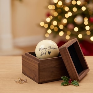 May include: A smooth, cream-colored spherical ornament featuring the black printed text Merry Christmas Dad with a small heart symbol, resting inside a square dark brown wooden box with an open lid, accompanied by a small gold snowflake charm.
