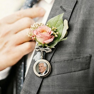Può includere: Una bottoniera floreale con una rosa rosa tenue, piccoli fiori bianchi di riempimento e foglie verdi fissata a una spilla in metallo argentato. Una cornice circolare argentata pende dalla spilla, contenente un ritratto in bianco e nero.