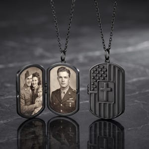 May include: A black metal dog tag locket pendant on a matching chain. The locket is shown open, revealing two sepia-toned photographs of people in military uniforms, and closed, displaying an embossed American flag design with a central cross.