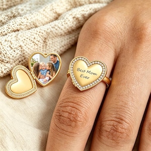 Könnte beinhalten: Ein goldener herzförmiger Ring mit der Aufschrift "Best Mom Ever" und einem Rand aus kleinen, klaren Steinen. Ein zweiter herzförmiger Anhänger öffnet sich und zeigt ein Familienfoto. Der Ring befindet sich an einem Finger.
