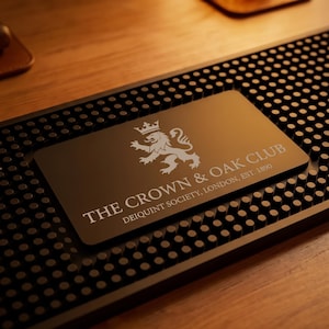May include: A black rectangular bar mat with a textured surface of small rubber nubs. Centered on the mat is a smooth black plaque featuring a white graphic of a crowned lion and the text THE CROWN & OAK CLUB DEIQUINT SOCIETY, LONDON, EST. 1890.