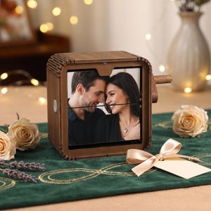 May include: A square wooden music box featuring a rotating display that shows a photograph of a couple. The dark wood frame has a textured, slatted design on the top and sides, with a small wooden crank handle extending from the right side.