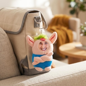 Pode incluir: Uma mochila de lona bege com acabamento cinza apresenta um chaveiro de pelúcia em forma de porco preso ao seu anel de metal. O porco de pelúcia tem um corpo rosa, cabelo verde brilhante, um nariz vermelho e uma camiseta azul com um rosto.