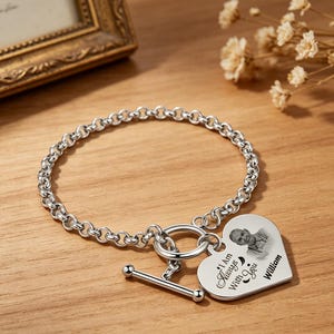 May include: A silver-toned chain bracelet featuring a toggle clasp and a heart-shaped pendant. The pendant is engraved with a portrait of a person, the text "I Am Always With You," and the name "William" in a black script and print font.