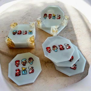 Peut inclure: Ensemble de dessous de verre en résine faits à la main, de couleur bleu clair. Chaque dessous de verre présente des personnages pixélisés rouges, bleus et blancs. Le support de dessous de verre a des accents de feuilles d'or. Un objet décoratif unique.