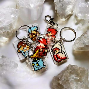 May include: A selection of pixel art character keyrings. Each keyring showcases a unique character design in bright colours, encased in clear, rectangular acrylic. The keyrings are attached to silver key rings and clasps.