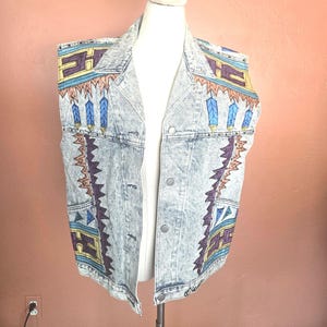 May include: A light blue acid-wash denim vest featuring a hand-painted geometric pattern in shades of purple, yellow, blue, and orange. The sleeveless garment has a classic collar, a button-front closure, and two front pockets with painted details.