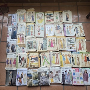 May include: A large collection of rectangular paper sewing pattern envelopes arranged in a grid. Each envelope features illustrations of clothing designs, including dresses, pants, and tops, with brand names like Butterick, Simplicity, and McCall's visible.