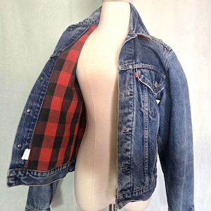 May include: A blue denim jacket with a red and black plaid flannel lining. The jacket has a classic design with button closures, pockets, and a collar. The jacket is displayed on a mannequin.