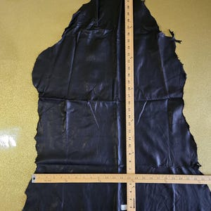 Könnte beinhalten: Ein großes, unregelmäßig geformtes Stück schwarzes Leder. Ein Holzlineal zeigt, dass das Leder etwa 89 cm lang ist. Das Leder hat eine strukturierte Oberfläche und wird vor einem goldenen Hintergrund gezeigt.